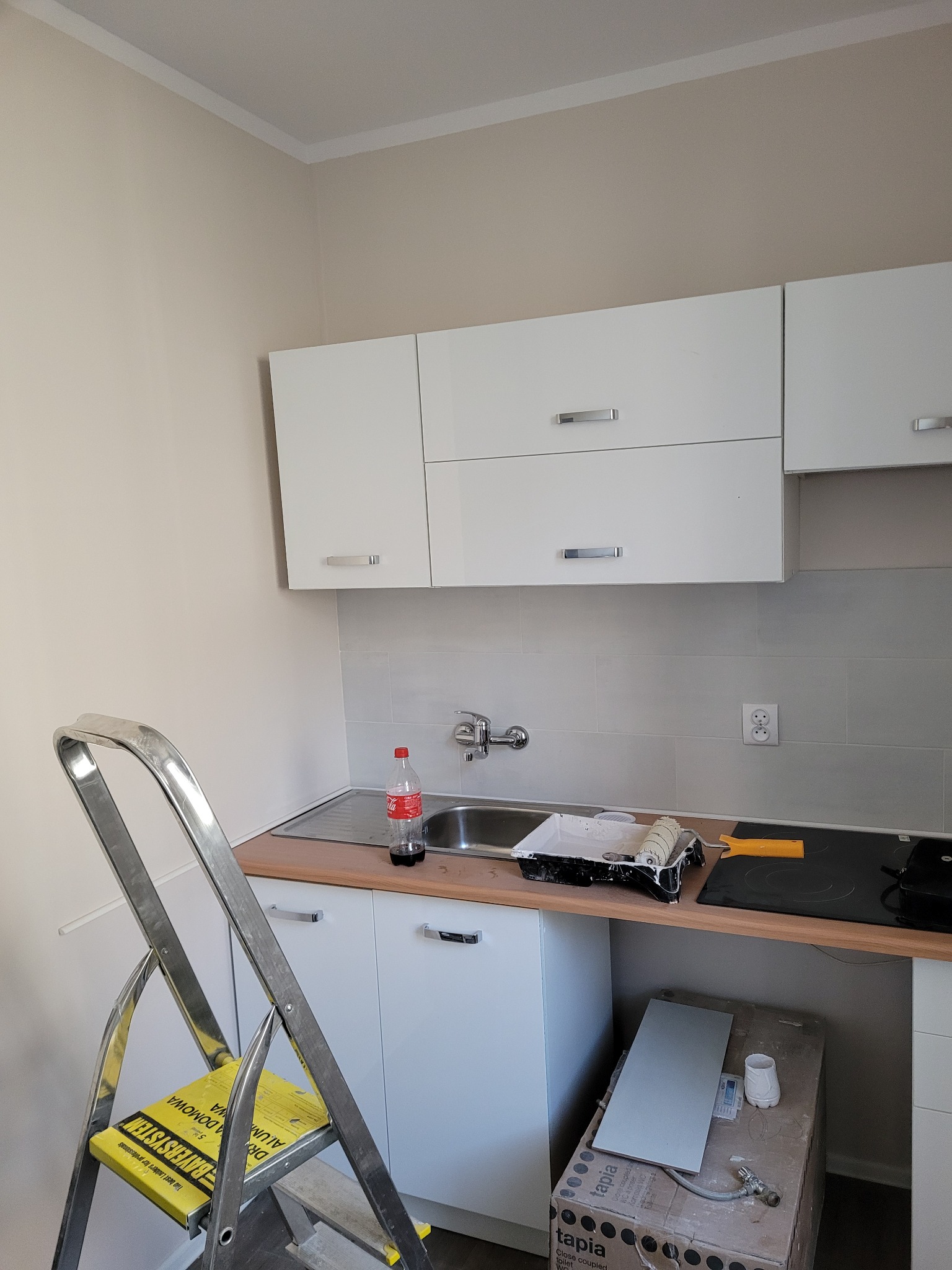Wnętrze kuchni w trakcie remontu: białe szafki, szare płytki na ścianie, aluminiowa drabina z żółtym siedziskiem, akcesoria malarskie i butelka napoju na blacie, karton z płytkami pod zlewem.