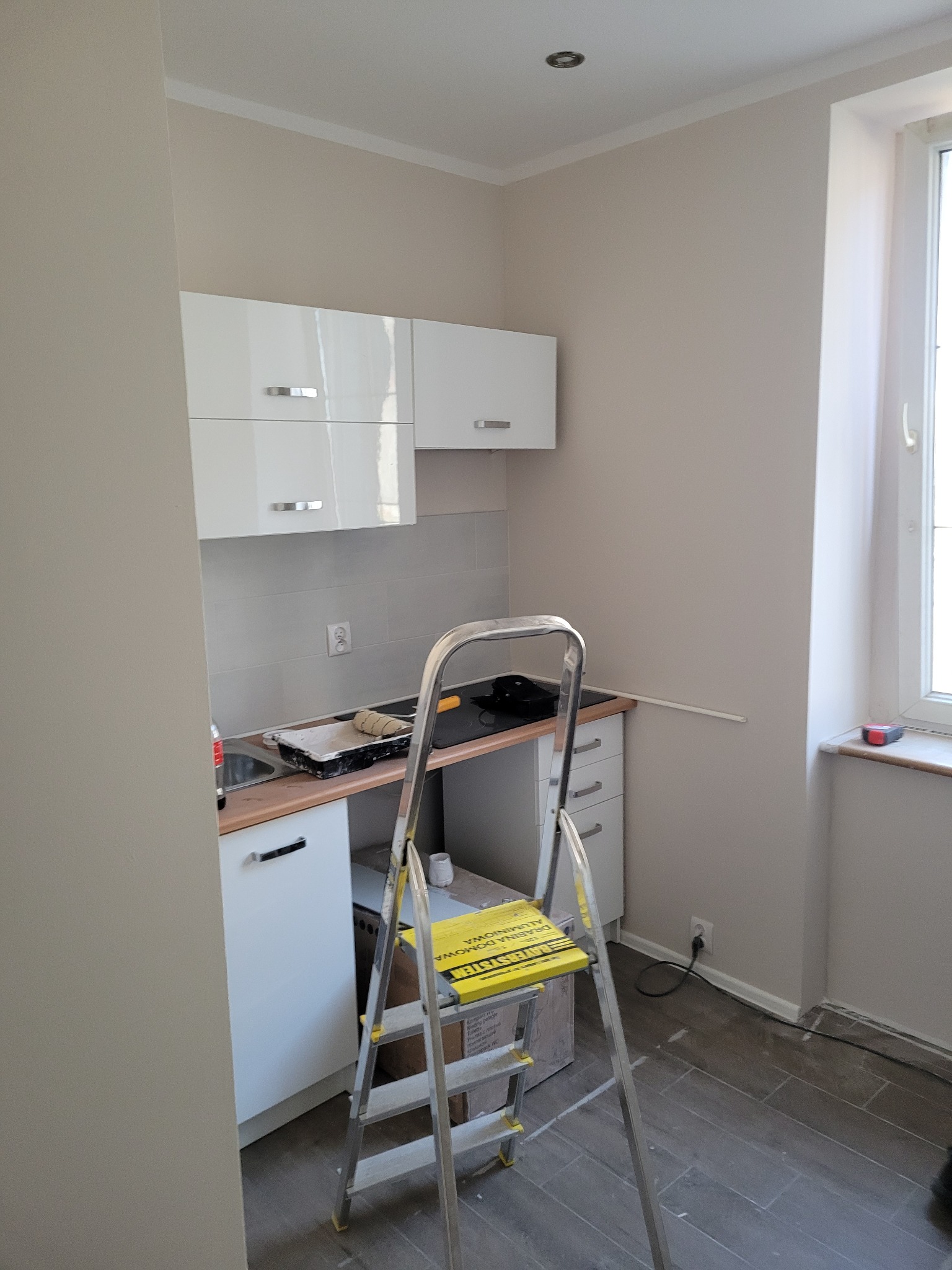 Wnętrze kuchni w trakcie malowania ścian na jasny beż, drabina aluminiowa stoi przed szafkami, widoczny wałek i kuweta z farbą na blacie.