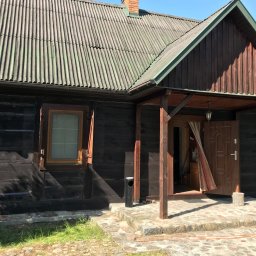 Agnes-Bud Agnieszka Figarska - Drewniany dom z szarozielonym dachem z eternitu, widoczne ślady mchu na dachu, ciemna elewacja z widocznymi słojami drewna, kamienny chodnik przed wejściem, podkowa nad oknem.