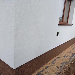 Agnes-Bud Agnieszka Figarska - Fragment otynkowanej na biało elewacji budynku z brązowym cokołem, widoczne okno z brązową ramą i fragment chodnika z piaskiem.
