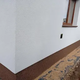 Fragment otynkowanej na biało elewacji budynku z brązowym cokołem, widoczne okno z brązową ramą i fragment chodnika z piaskiem.