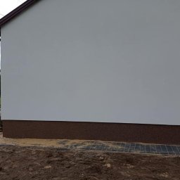 Agnes-Bud Agnieszka Figarska - Fragment bocznej ściany budynku z białą elewacją i ciemnobrązowym cokołem, widoczny fragment okna z firanką, obok ułożona szara kostka brukowa, a dalej ziemia i trawa, w tle pole i słupy energetyczne.