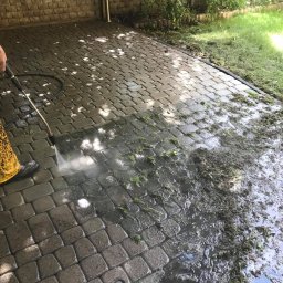 Agnes-Bud Agnieszka Figarska - Czyszczenie kostki brukowej z resztkami trawy za pomocą myjki ciśnieniowej; widoczna różnica między czystą a brudną powierzchnią, osoba w żółtych spodniobutach.