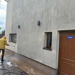 Agnes-Bud Agnieszka Figarska - Pracownik w żółtym płaszczu czyści elewację budynku myjką ciśnieniową. Widoczne różnice w czystości na ścianie i kostce brukowej. W tle okno i drzwi z napisem 'Wejście'.