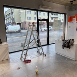 Agnes-Bud Agnieszka Figarska - Wnętrze lokalu z aluminiową drabiną stojącą przed dużym oknem, widoczne ślady po czyszczeniu, butelka ze środkiem czyszczącym na podłodze. Widok na ulicę przez okno.