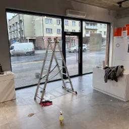 Wnętrze lokalu z aluminiową drabiną stojącą przed dużym oknem, widoczne ślady po czyszczeniu, butelka ze środkiem czyszczącym na podłodze. Widok na ulicę przez okno.