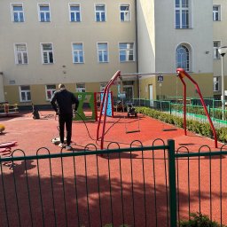 Agnes-Bud Agnieszka Figarska - Pracownik w czarnej kurtce czyści czerwoną nawierzchnię placu zabaw za pomocą specjalistycznej maszyny. W tle budynek z licznymi oknami.