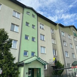 Agnes-Bud Agnieszka Figarska - Pracownik na podnośniku czyści elewację bloku. Widoczna różnica w kolorze między umytą a brudną częścią. Na dole rowery i maszyna czyszcząca.