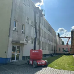 Częściowo umyta elewacja budynku z podnośnika Manitou. Na drugim planie kolejny podnośnik. Widoczne okna i trawnik. Słoneczny dzień.