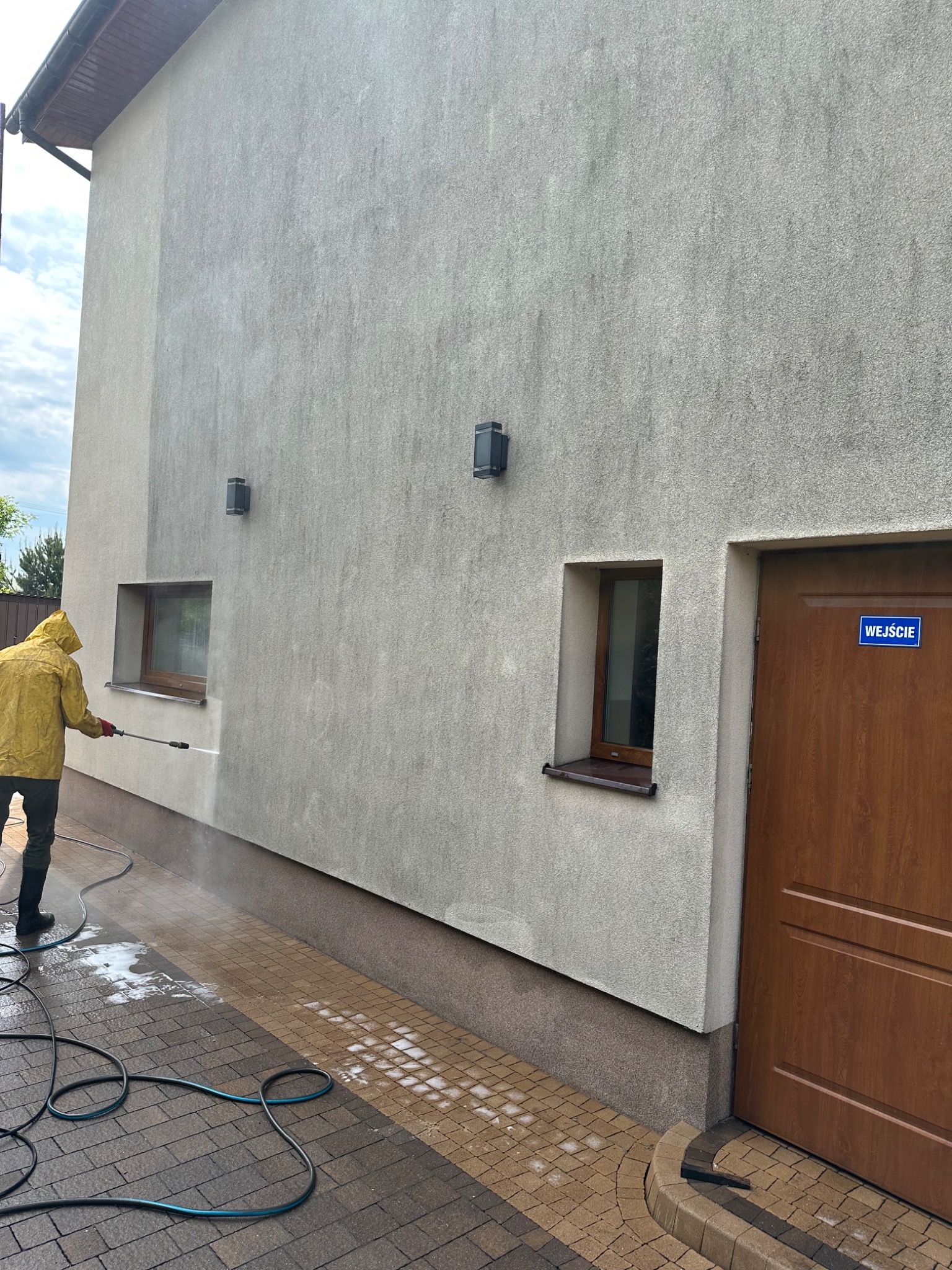 Pracownik w żółtym płaszczu czyści elewację budynku myjką ciśnieniową. Widoczne różnice w czystości na ścianie i kostce brukowej. W tle okno i drzwi z napisem 'Wejście'.