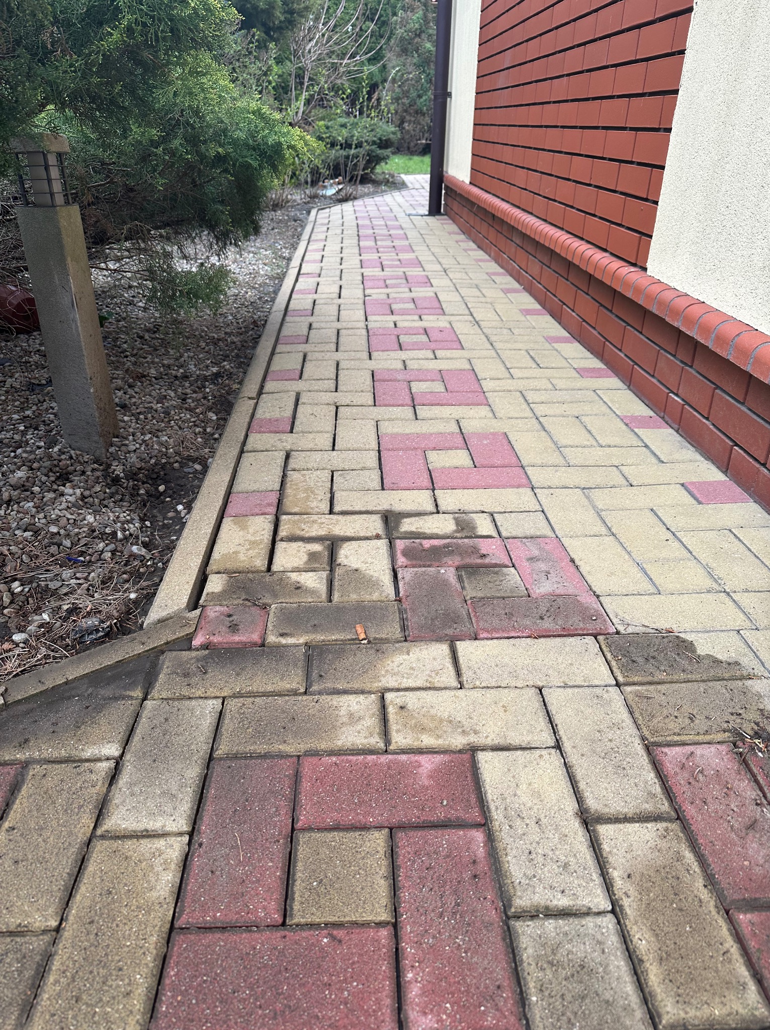 Chodnik z kostki brukowej w odcieniach beżu i czerwieni prowadzący wzdłuż budynku z ceglaną elewacją. Widoczne zabrudzenia na kostce i fragment roślinności.