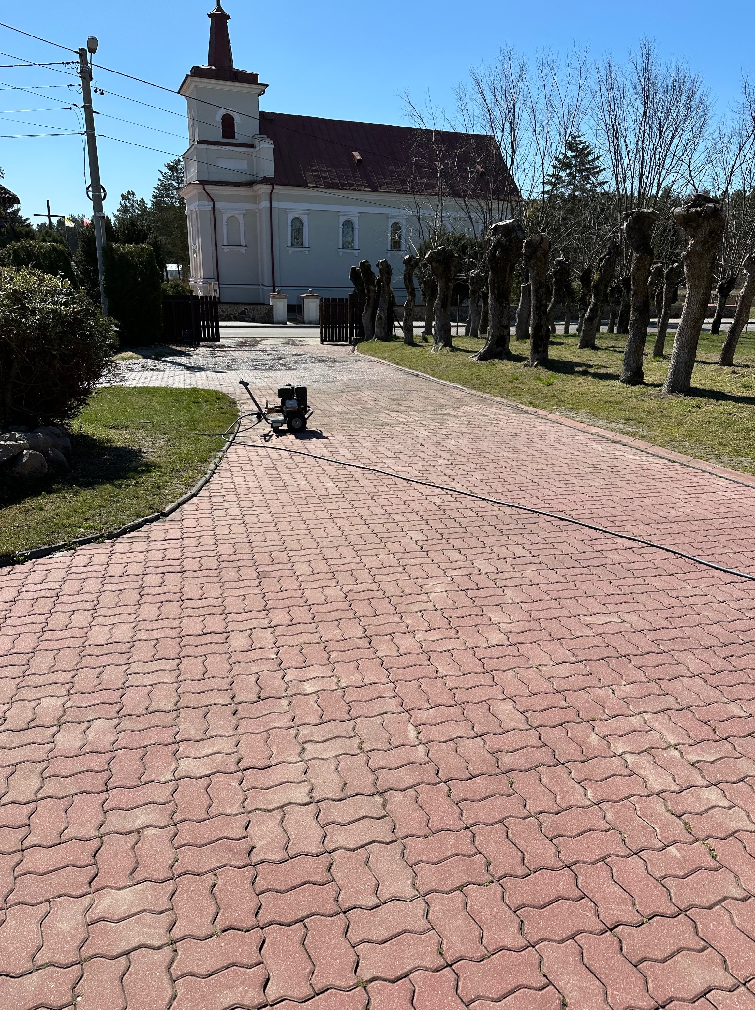 Czerwona kostka brukowa na podjeździe, sprzęt do czyszczenia z wężem, w tle kościół i drzewa. Słoneczny dzień.