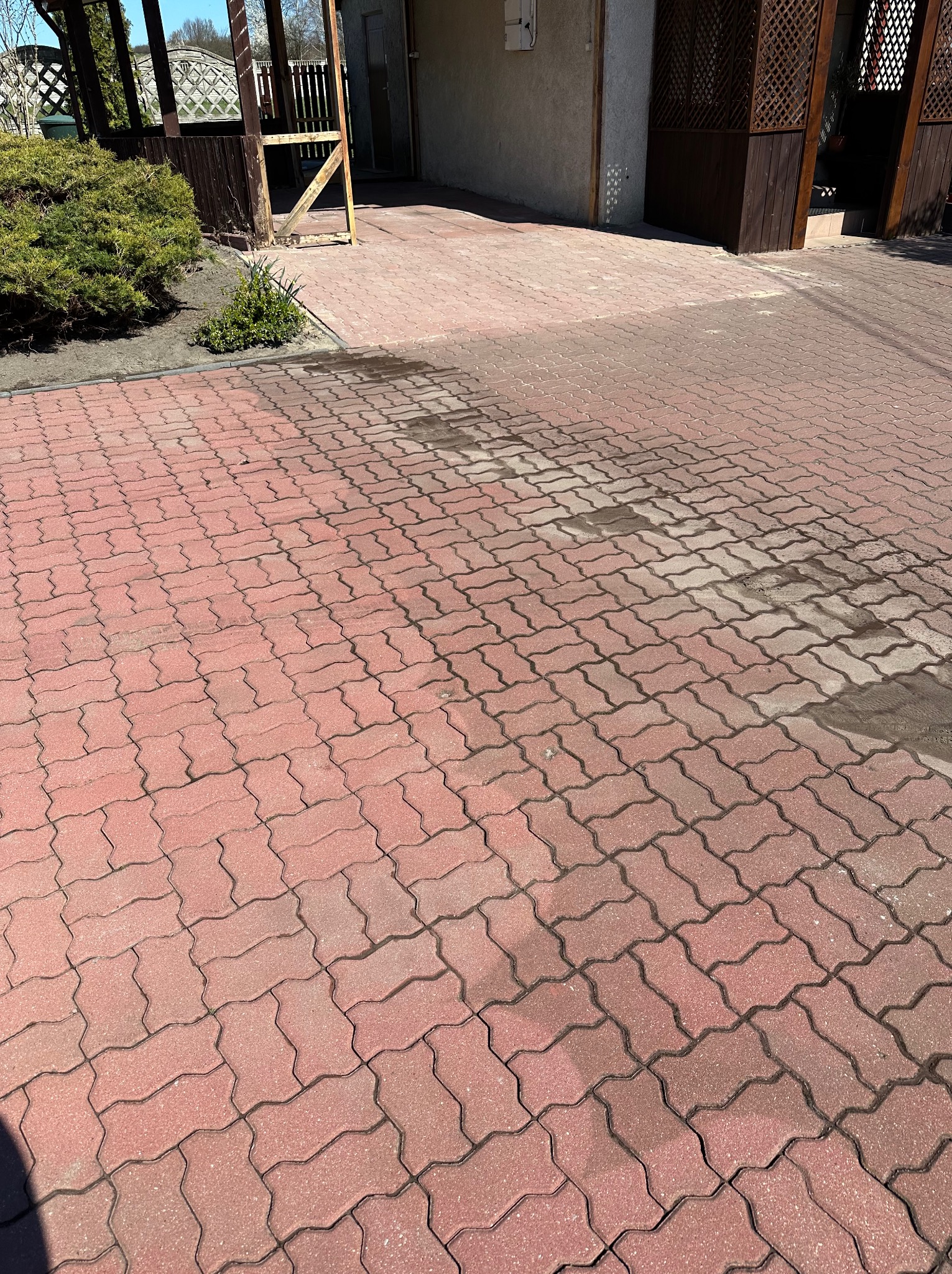 Czerwona kostka brukowa ułożona na podjeździe, częściowo mokra, sugerująca niedawne mycie. Widoczny fragment budynku i elementy architektury ogrodowej.