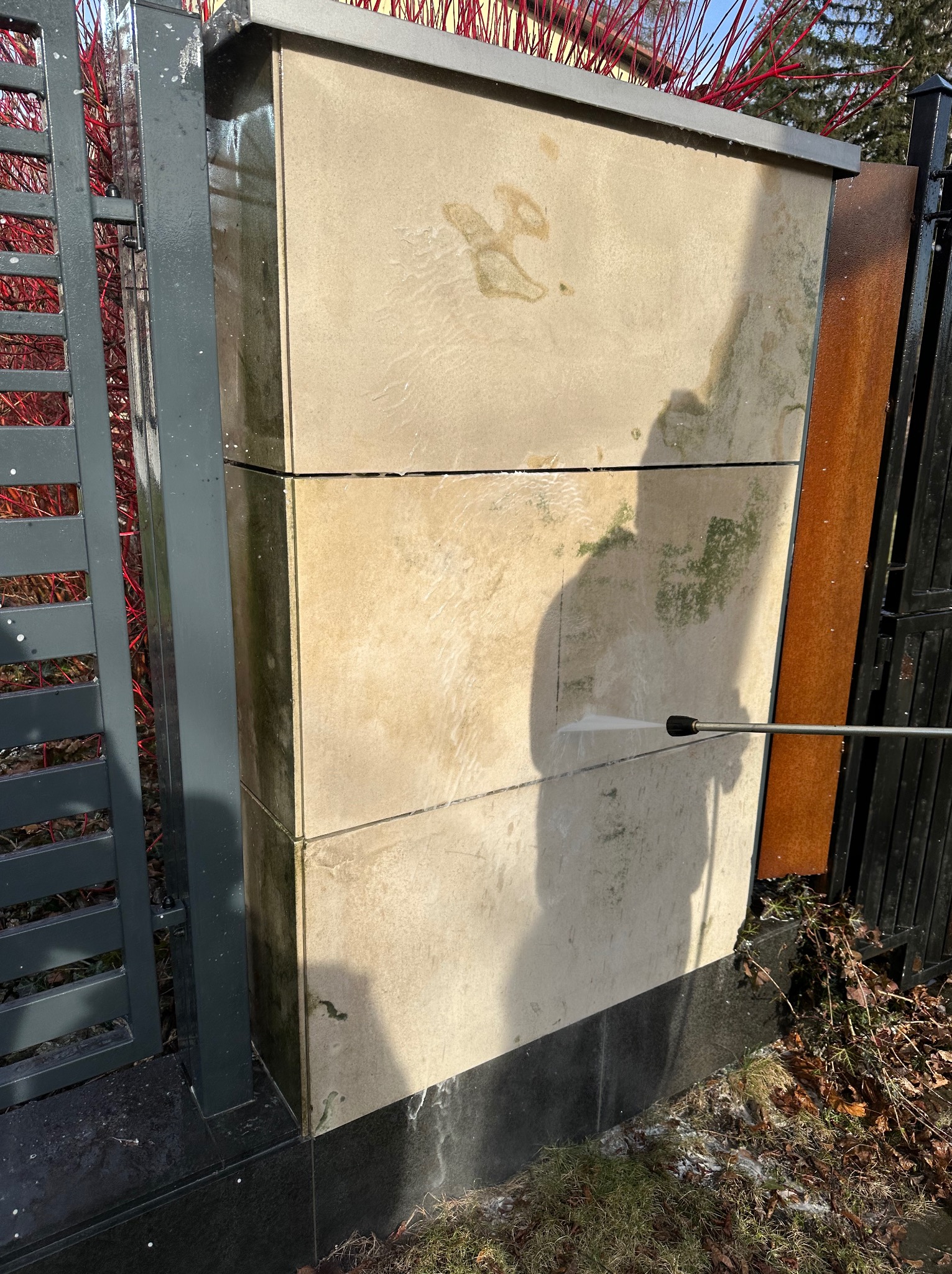 Czyszczenie elewacji z kamienia za pomocą myjki ciśnieniowej. Widoczne zabrudzenia i efekt czyszczenia strumieniem wody. Metalowe ogrodzenie w tle.