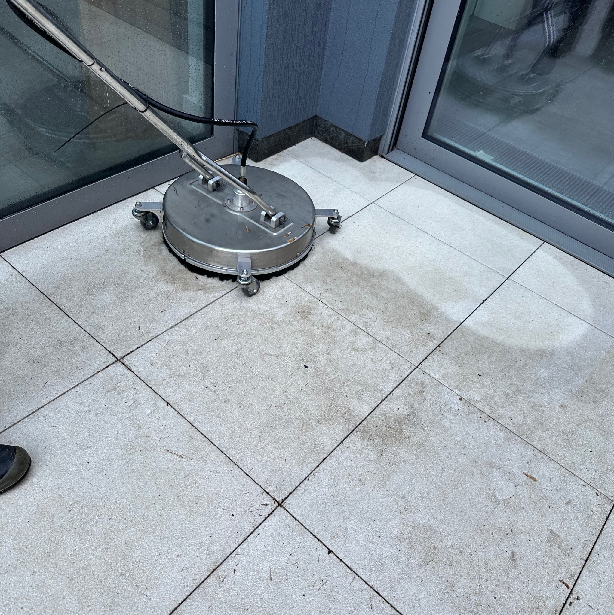 Mycie kostki brukowej przy użyciu myjki ciśnieniowej z okrągłą dyszą. Widoczny fragment czystej kostki i fragment buta operatora. Praca w toku.