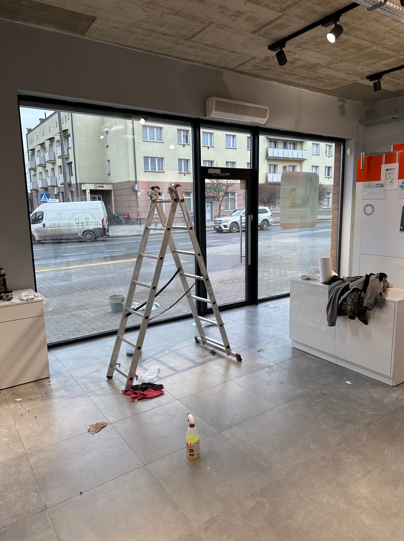 Wnętrze lokalu z aluminiową drabiną stojącą przed dużym oknem, widoczne ślady po czyszczeniu, butelka ze środkiem czyszczącym na podłodze. Widok na ulicę przez okno.