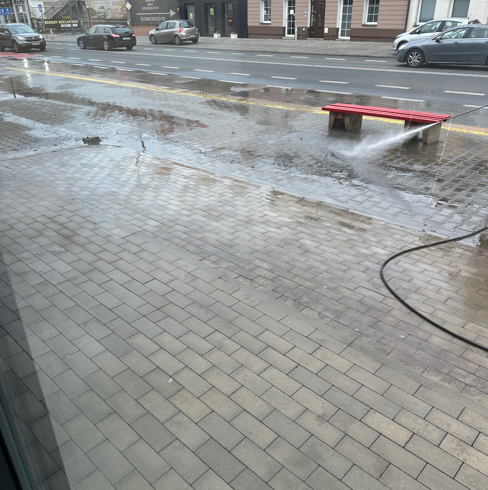 Czyszczenie kostki brukowej przy ulicy za pomocą myjki ciśnieniowej. Czerwona ławka obok. Widoczne mokre i suche fragmenty kostki. Ulica z samochodami w tle.