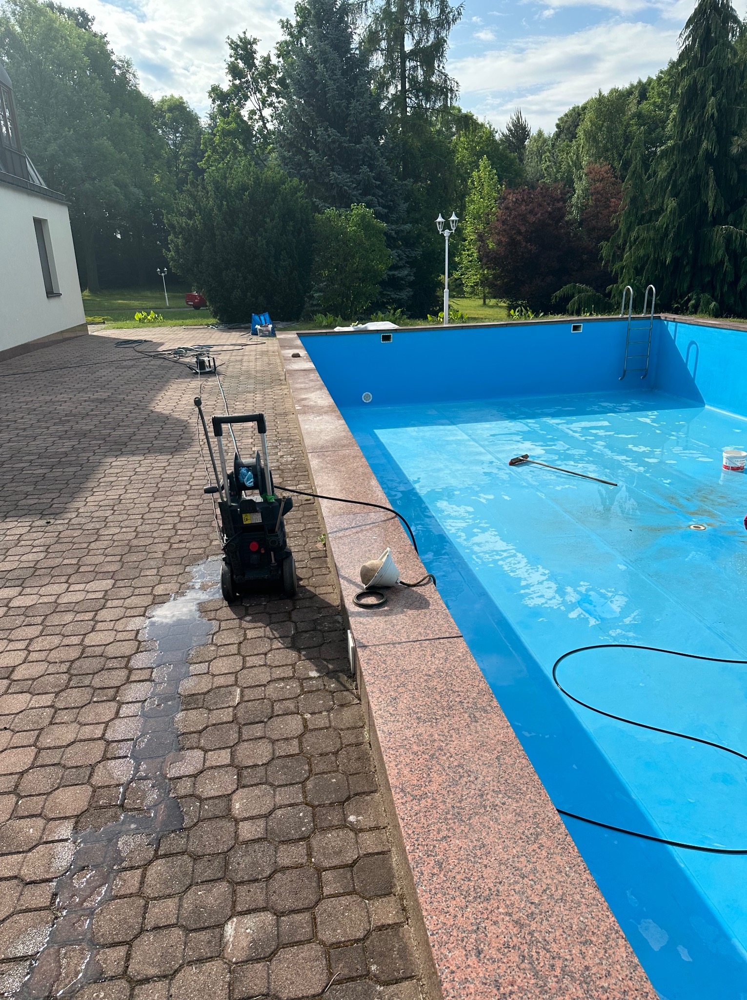 Czyszczenie basenu zewnętrznego z użyciem myjki ciśnieniowej. Widoczne ślady czyszczenia na kostce brukowej i wewnątrz niecki basenowej. Sezonowe przygotowanie.