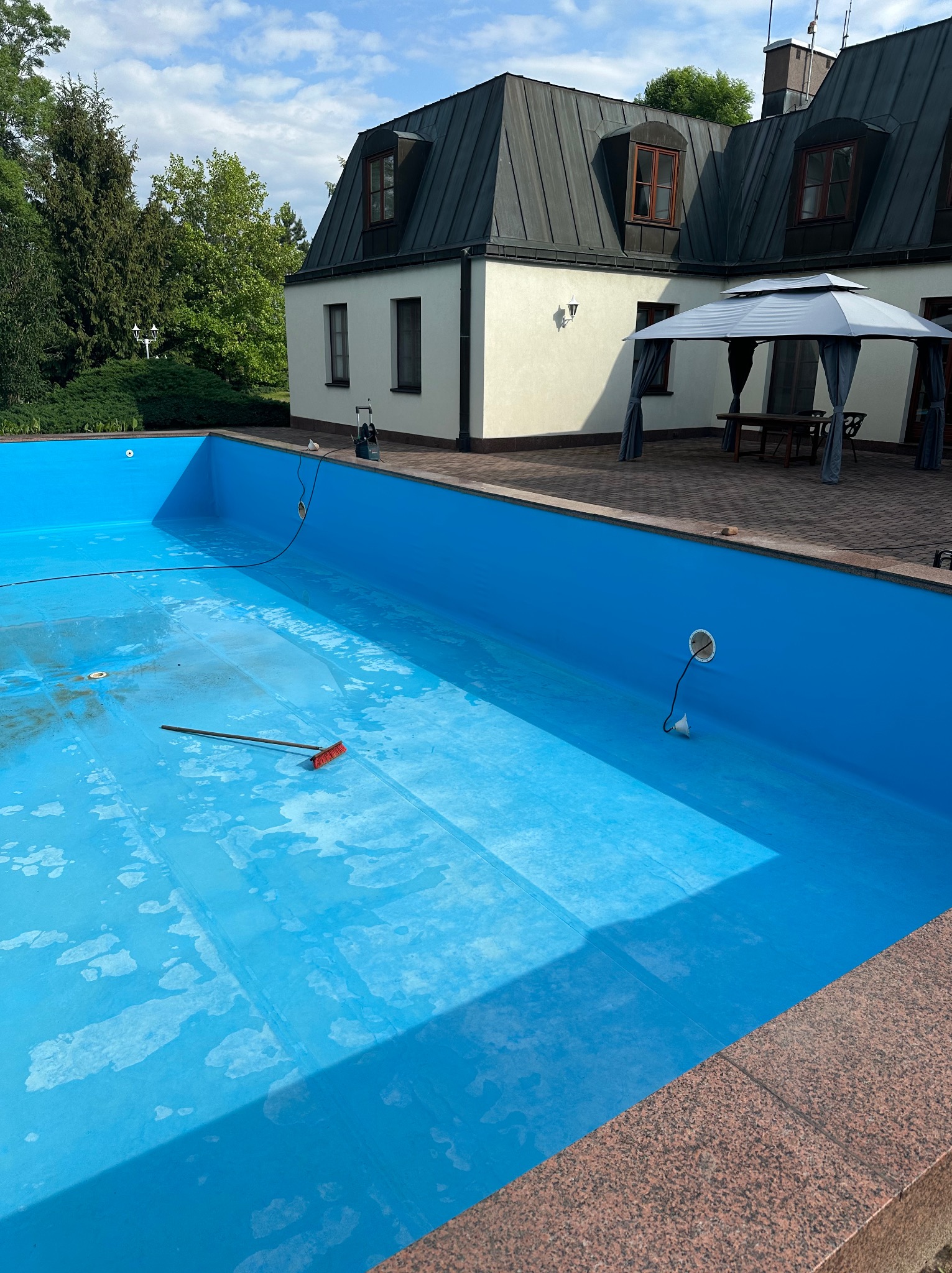 Pusty, wyłożony błękitną folią basen ogrodowy przed czyszczeniem. Wewnątrz leży szczotka, w tle dom z ciemnym dachem i altana.