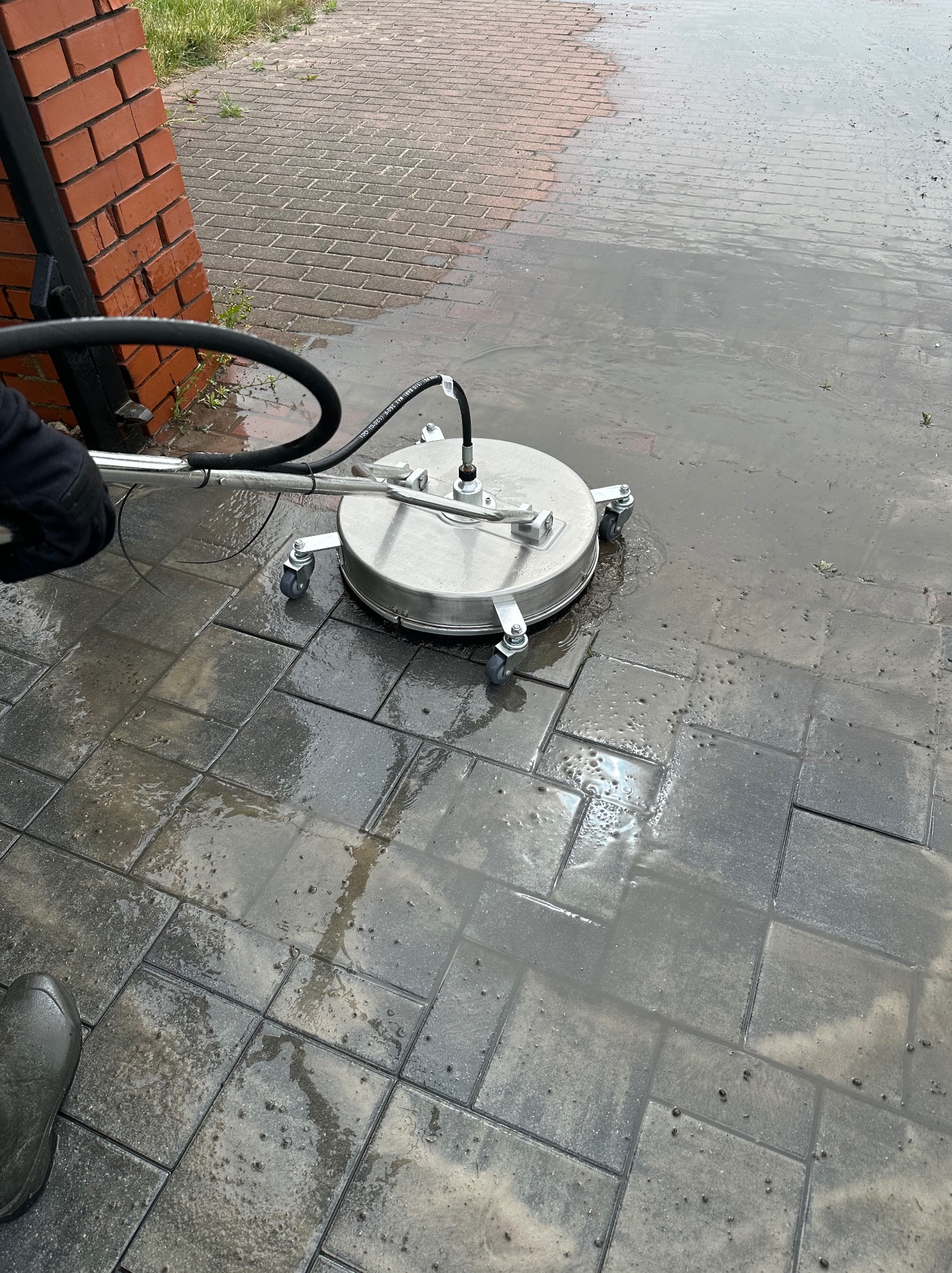 Czyszczenie kostki brukowej myjką ciśnieniową z okrągłą dyszą. Widoczne mokre, szare kostki i fragment węża doprowadzającego wodę. Praca w toku.