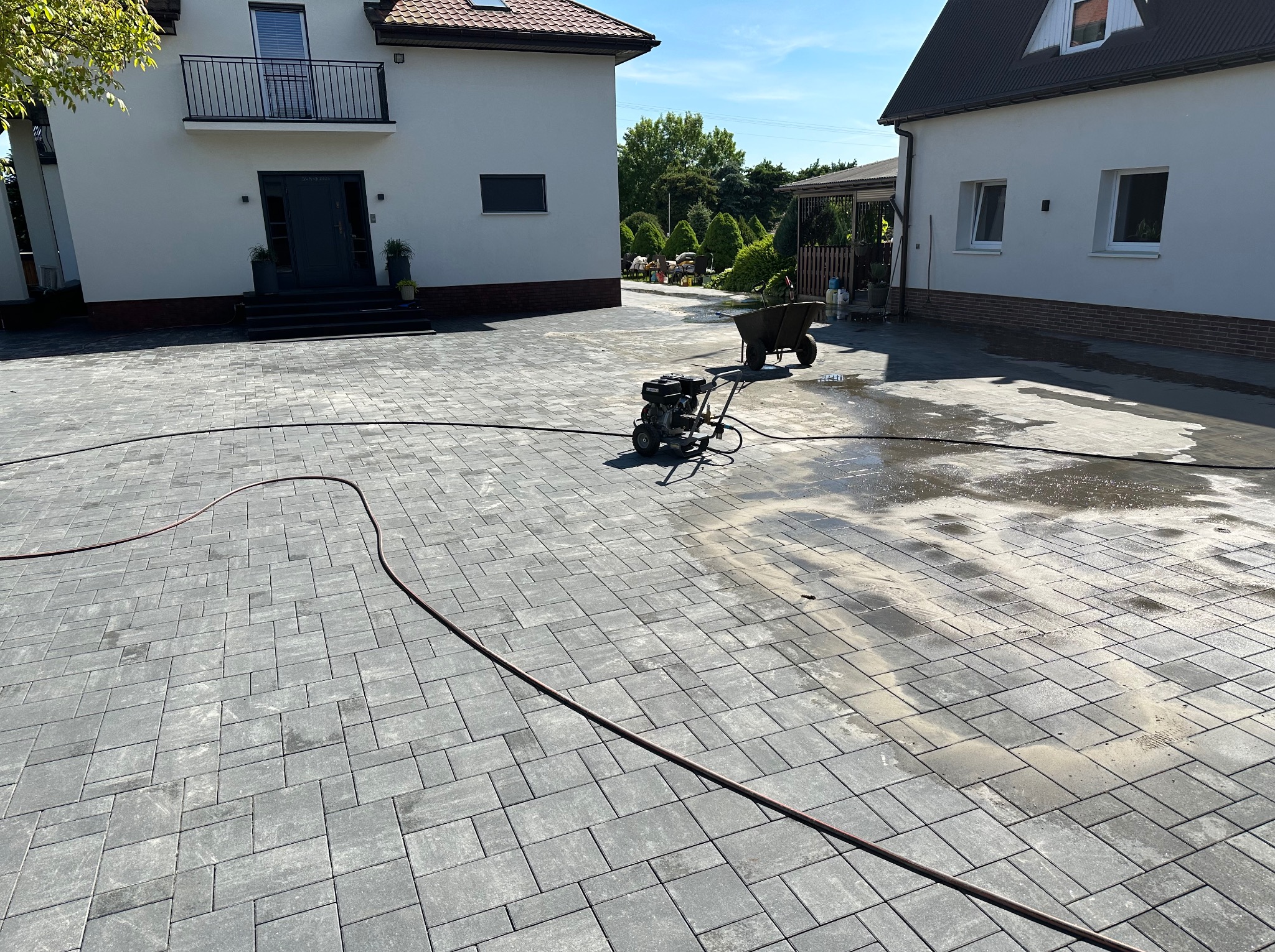 Czyszczenie kostki brukowej myjką ciśnieniową na posesji z dwoma budynkami, widoczne mokre i suche fragmenty kostki oraz wąż.