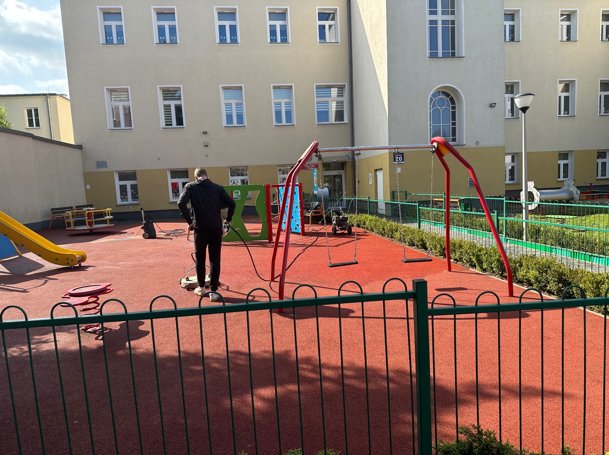 Pracownik w czarnej kurtce czyści czerwoną nawierzchnię placu zabaw za pomocą specjalistycznej maszyny. W tle budynek z licznymi oknami.