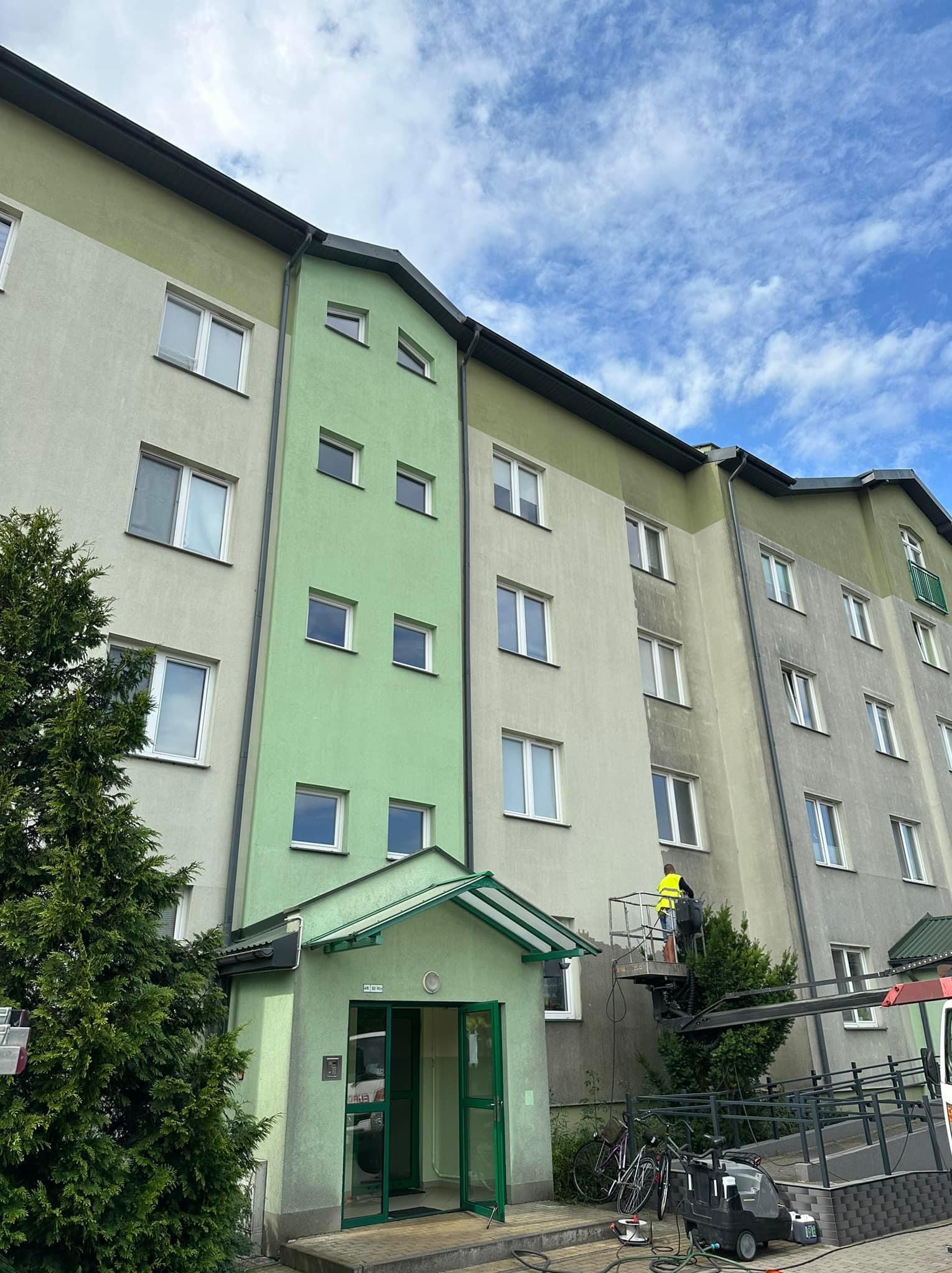 Pracownik na podnośniku czyści elewację bloku. Widoczna różnica w kolorze między umytą a brudną częścią. Na dole rowery i maszyna czyszcząca.