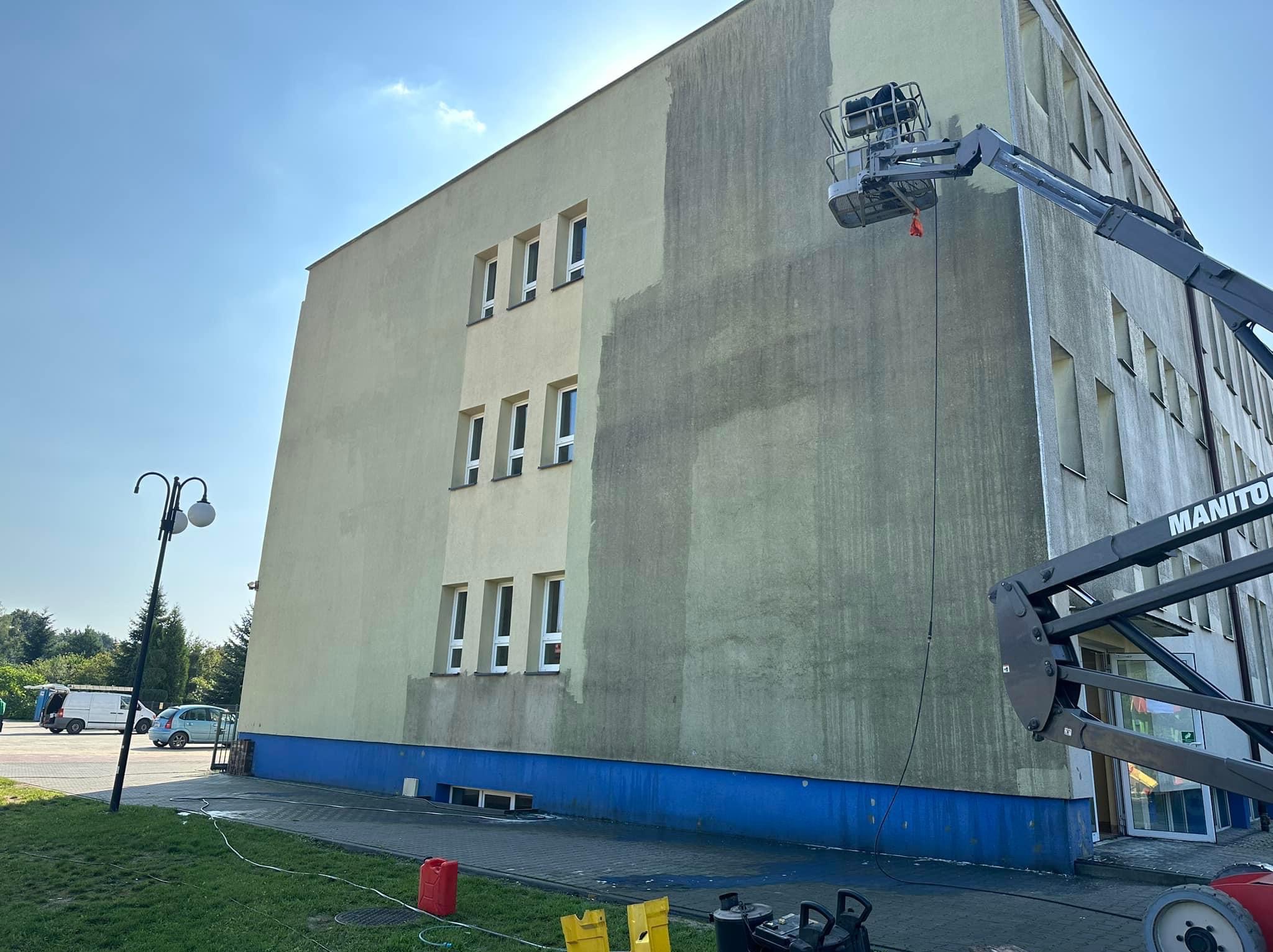 Częściowo umyta elewacja budynku z podnośnika koszowego. Widoczne różnice w kolorze elewacji przed i po czyszczeniu. Na dole widoczne urządzenia do mycia.