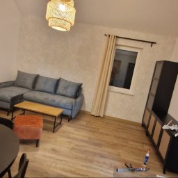 DeBest Wykończenia Wnętrz Remonty - Jasne wnętrze pokoju dziennego z szarą sofą narożną, pomarańczowym fotelem, okrągłym stołem i czarną szafką RTV; na podłodze widoczne narzędzia budowlane.