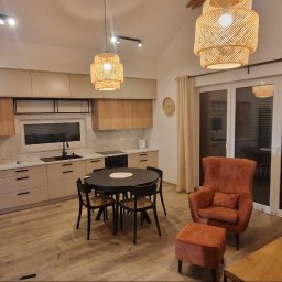 DeBest Wykończenia Wnętrz Remonty - Nowoczesna kuchnia z beżowymi szafkami, czarnym sprzętem AGD i okrągłym stołem jadalnym, uzupełniona o wiklinowe lampy wiszące i fotel z podnóżkiem w odcieniach pomarańczy.