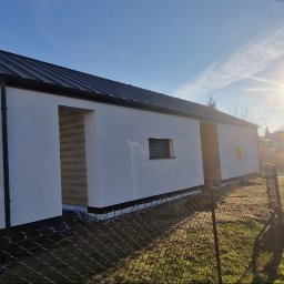 DeBest Wykończenia Wnętrz Remonty - Nowoczesny, parterowy budynek z czarnym, metalowym dachem i białymi ścianami zewnętrznymi w surowym stanie, widoczny przez ogrodzenie z siatki, w tle błękitne niebo i ostre słońce.