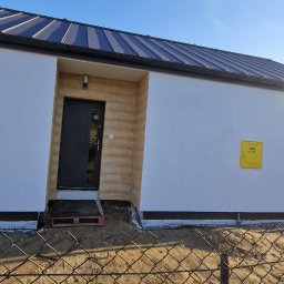 DeBest Wykończenia Wnętrz Remonty - Nowoczesny, jednokondygnacyjny dom z czarnym dachem i drzwiami wejściowymi, widoczna żółta skrzynka gazowa na białej ścianie, ujęcie zza ogrodzenia z siatki.