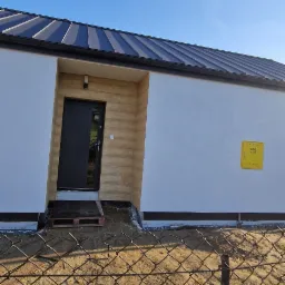 Nowoczesny, jednokondygnacyjny dom z czarnym dachem i drzwiami wejściowymi, widoczna żółta skrzynka gazowa na białej ścianie, ujęcie zza ogrodzenia z siatki.