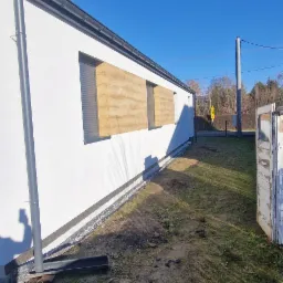 Elewacja budynku jednorodzinnego po remoncie, z widocznym ociepleniem i nową strukturą tynku na fragmentach ściany, rynna spustowa, teren wokół budynku częściowo zagospodarowany, błękitne niebo.