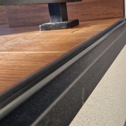 DeBest Wykończenia Wnętrz Remonty - Szczegółowe ujęcie zabudowy balkonu z drewnopodobną podłogą, czarną ramą i oświetloną fakturą ściany.