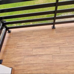 DeBest Wykończenia Wnętrz Remonty - Balkon z nową podłogą z płytek imitujących drewno, czarna metalowa balustrada, pomarańczowa kuweta z resztkami zaprawy i butelka po środku czyszczącym