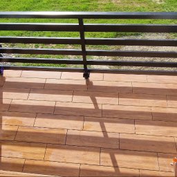 DeBest Wykończenia Wnętrz Remonty - Balkon z drewnopodobną terakotą i czarną, metalową balustradą, widok z góry, fragment samochodu w tle.