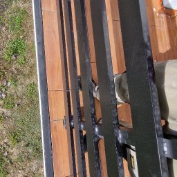 DeBest Wykończenia Wnętrz Remonty - Wysoki kąt ujęcia na montaż czarnej metalowej konstrukcji na balkonie wyłożonym płytkami imitującymi drewno, widoczne narzędzia i fragment buta montera.