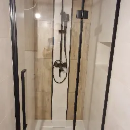 Kabina prysznicowa z czarnymi profilami i deszczownicą, obudowana jasnymi płytkami imitującymi drewno i białymi płytkami.