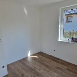 Pusty, pomalowany na biało pokój z brązowymi panelami podłogowymi, przewody wystające z sufitu i materiały budowlane na parapecie.