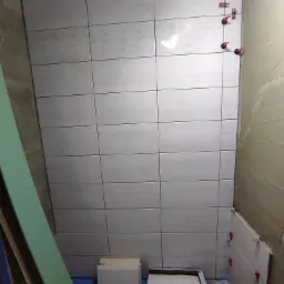 Układanie jasnoszarych płytek z delikatnym wzorem na ścianie w łazience, widoczne czerwone klipsy poziomujące, surowy tynk na sąsiedniej ścianie, karton z płytkami na podłodze.