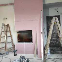 Wnętrze w trakcie tworzenia obudowy kominka z płyt kartonowo-gipsowych, widoczne dwie drewniane drabiny, poziomica, wkrętarka i nieporządek budowlany.