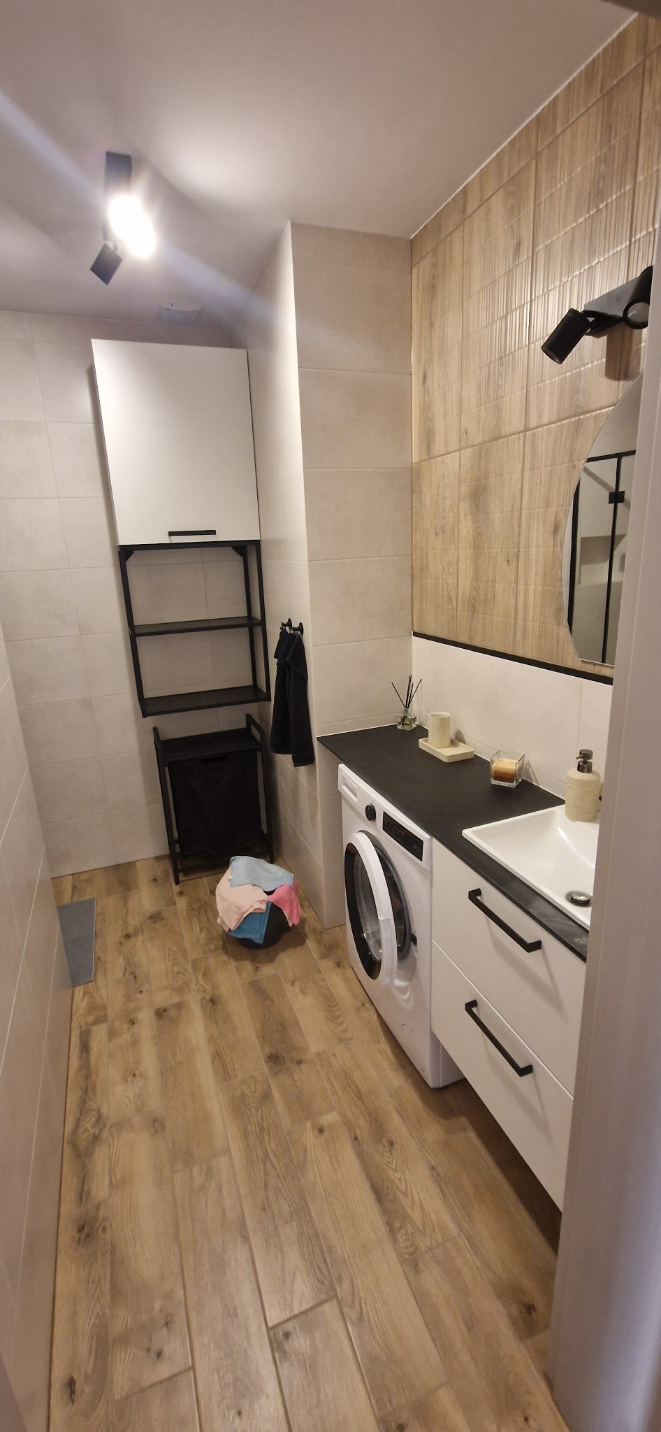 Umywalka z szafką i pralka pod blatem w nowoczesnej łazience z drewnopodobnymi płytkami, koszem na pranie i wiszącą szafką.