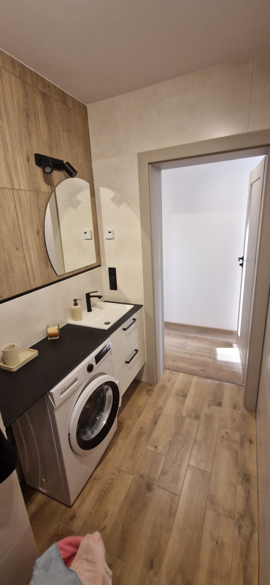 Nowoczesna łazienka z pralką zabudowaną pod blatem umywalki, drewnopodobne płytki na podłodze i ścianie, czarne akcenty w armaturze i oświetleniu, widok na otwarte drzwi do innego pomieszczenia.