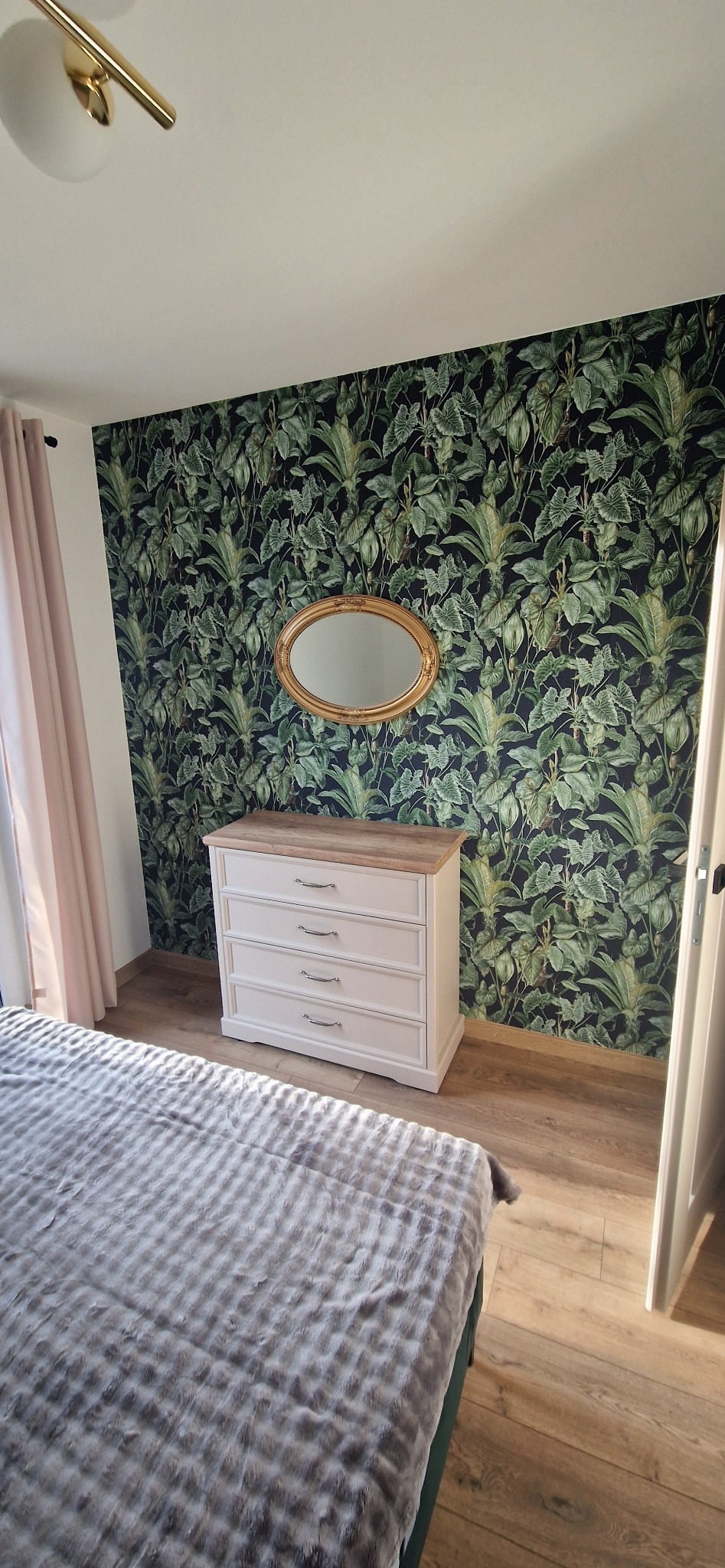 Pokój z komodą z czterema szufladami, lustrem w złotej ramie i tapetą w tropikalne liście na ścianie, widok na łóżko z szarą narzutą.