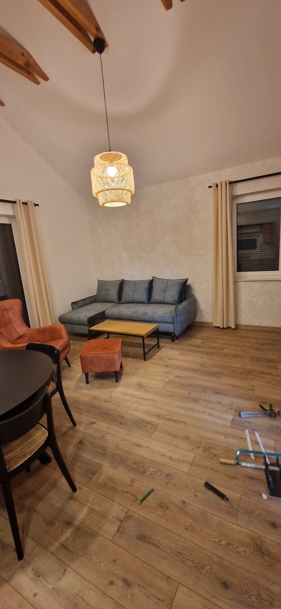 Jasne wnętrze po remoncie z drewnopodobną podłogą, szarą sofą, okrągłym stołem z krzesłami i wiszącą lampą z naturalnego materiału, widoczne narzędzia na podłodze sugerują prace wykończeniowe.