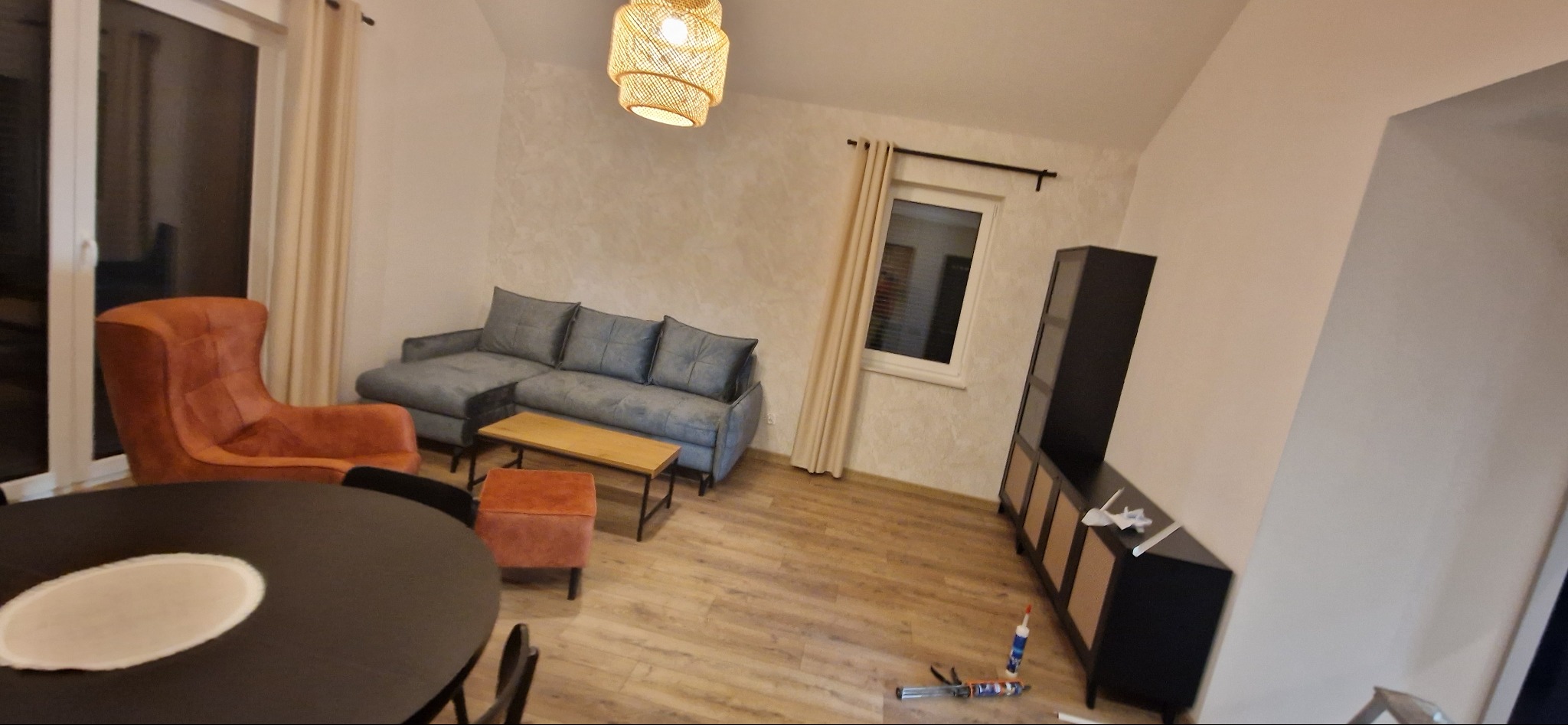 Jasne wnętrze pokoju dziennego z szarą sofą narożną, pomarańczowym fotelem, okrągłym stołem i czarną szafką RTV; na podłodze widoczne narzędzia budowlane.