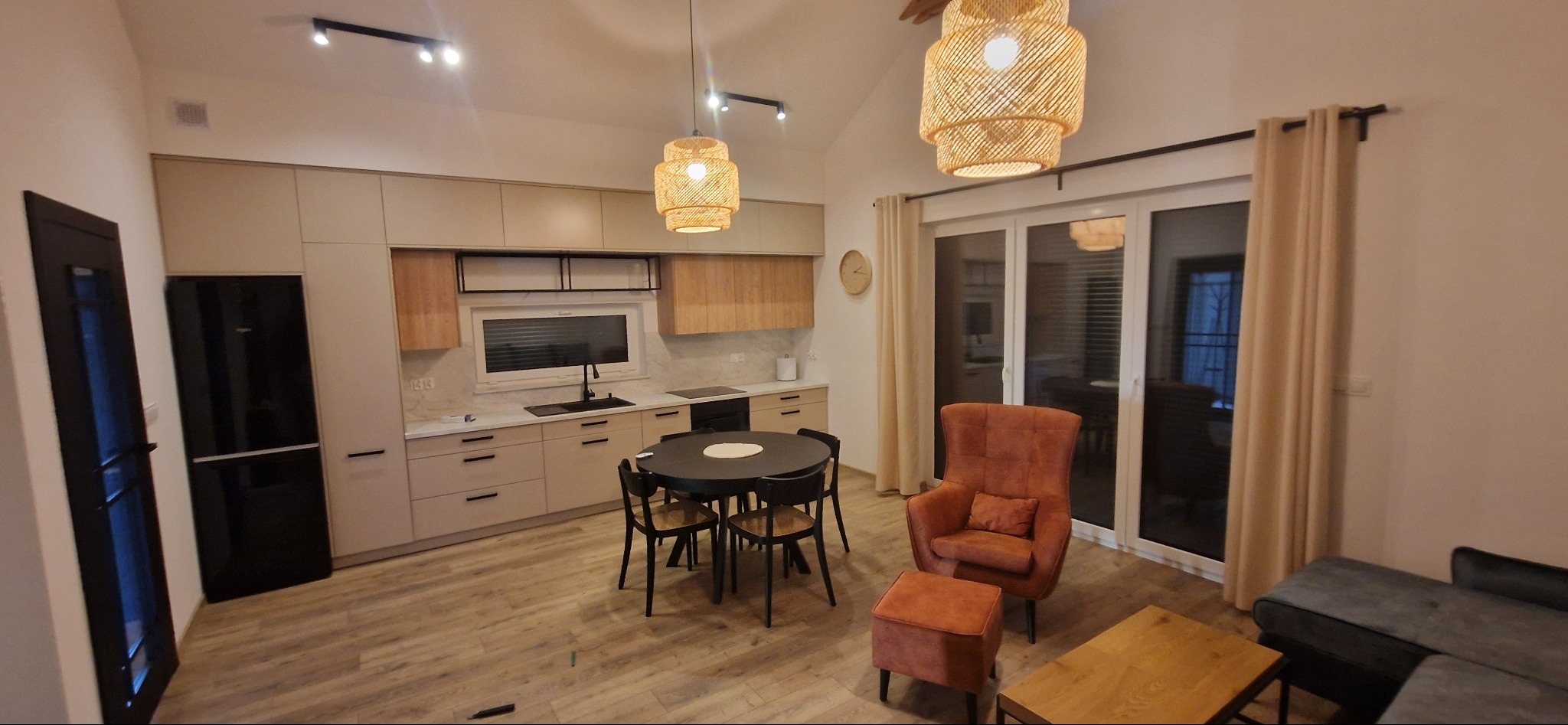 Nowoczesna kuchnia z beżowymi szafkami, czarnym sprzętem AGD i okrągłym stołem jadalnym, uzupełniona o wiklinowe lampy wiszące i fotel z podnóżkiem w odcieniach pomarańczy.