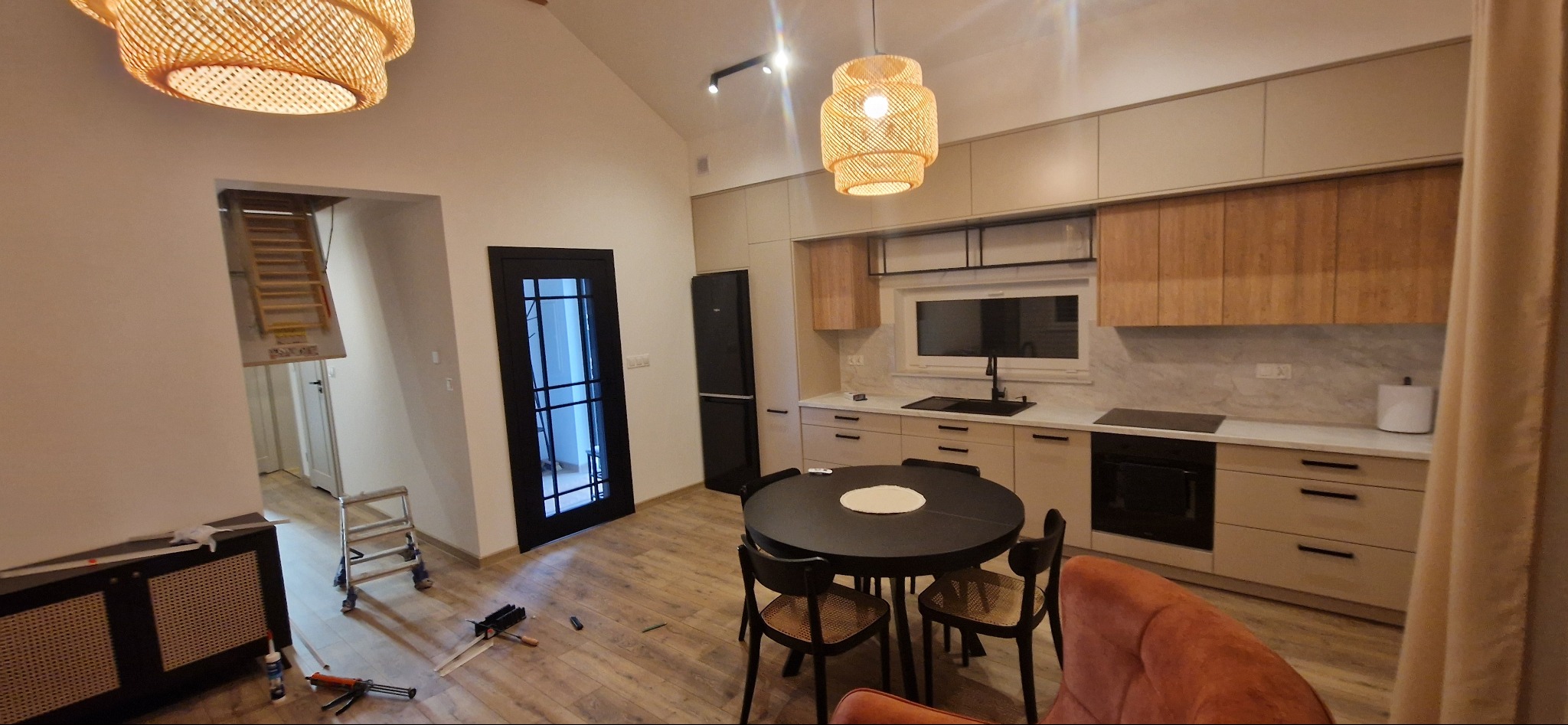 Nowoczesna kuchnia z drewnianymi i beżowymi szafkami, czarnym stołem i krzesłami, oraz lampami z abażurami z naturalnego materiału. W tle widoczna czarna lodówka i czarne drzwi z szybami.