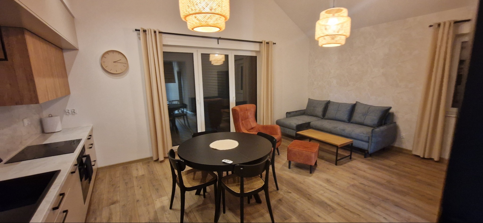 Jasne wnętrze mieszkania z drewnianą podłogą, okrągłym stołem z rattanowymi krzesłami, szarą sofą, pomarańczowym fotelem i lampami z naturalnego materiału